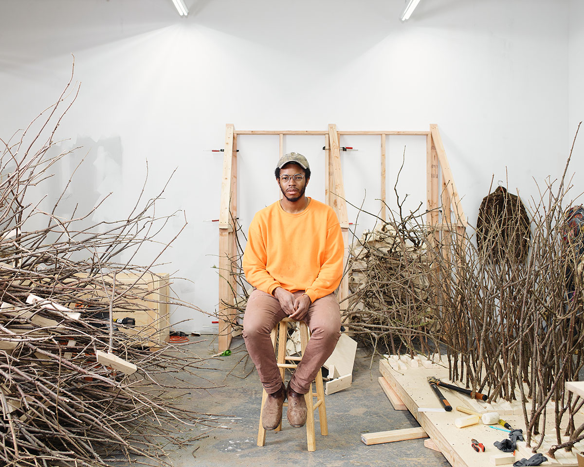 A black man in a yellow shirt and tan pants sitting on a stool surrounded by wood sculptural artworks
