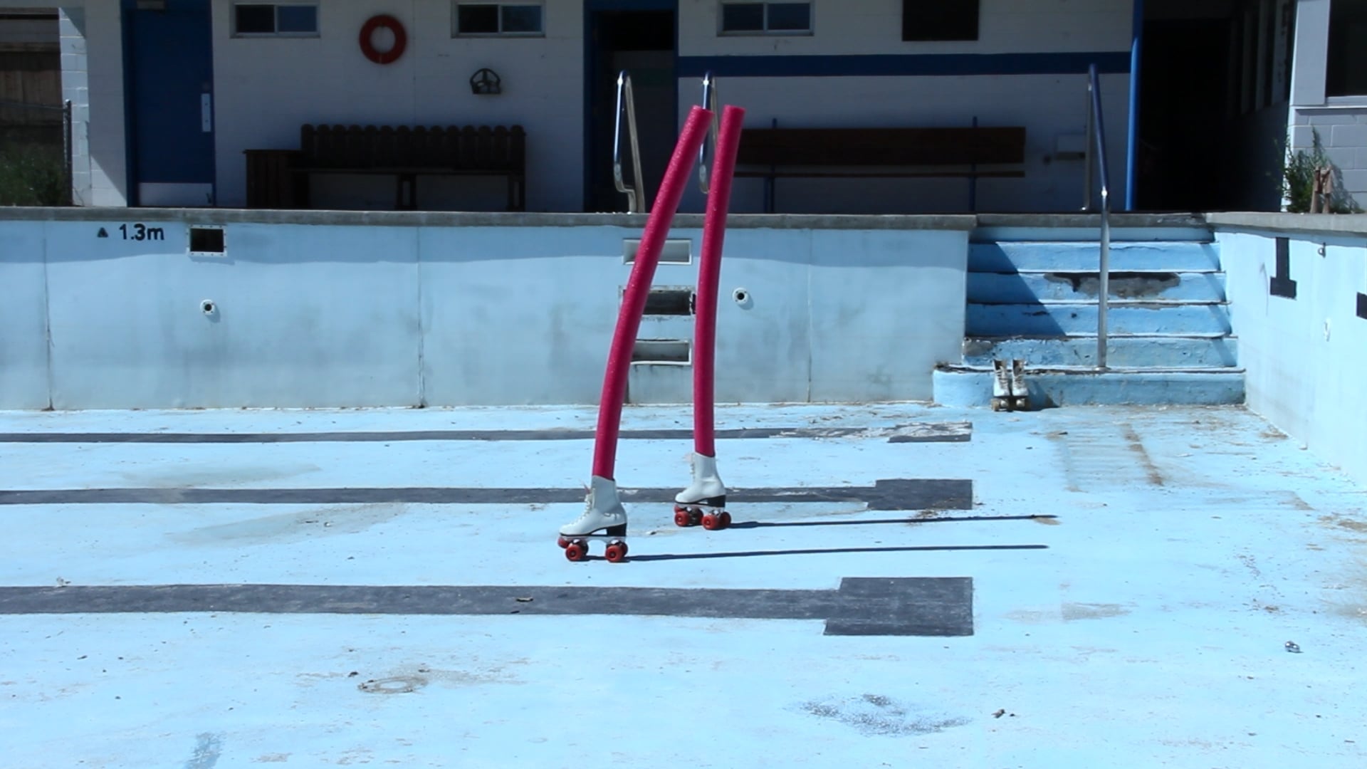 An still from a video of pool noodles stuck vertically into roller skates in an empty pool